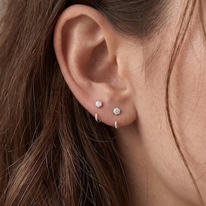 May include: Two silver earrings are worn in the earlobe. Each earring features a small, clear, round-cut gemstone set in a prong setting, paired with a simple, thin, silver U-shaped hoop that hangs directly below the gemstone stud.