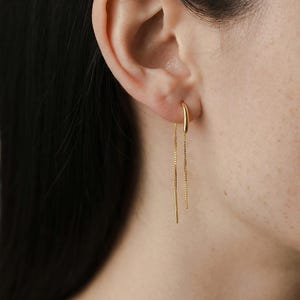 May include: A close-up of a gold earring. The earring features a small hoop that connects to a delicate chain. A gold bar hangs from the chain. The earring is worn in the ear of a person.