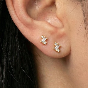 Könnte beinhalten: Nahaufnahme eines Ohrs mit zwei kleinen, goldfarbenen Ohrsteckern. Jeder Ohrring hat einen rechteckigen, klaren Edelstein, der von kleineren, runden, klaren Edelsteinen flankiert wird. Die Ohrringe sind auf der Haut.