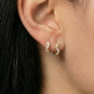 May include: Close-up of a person's ear adorned with two small, gold-colored hoop earrings. Each earring features a series of clear, sparkling gemstones arranged in a curved, zigzag pattern. The earrings are set against the backdrop of the ear and dark hair.