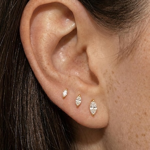 May include: Close-up of an ear with three gold stud earrings. The earrings feature marquise-cut diamonds, each set in a gold bezel. The ear has freckles and dark hair is visible.