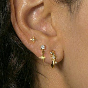 May include: A selection of gold earrings, including a star stud, a dangling star and square gem, and a small hoop with star and gem details. The earrings are displayed on an ear.