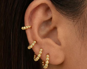 May include: Gold-colored beaded hoop earrings and ear cuff. The earrings are small, with a series of tiny gold beads forming a continuous circle. The ear cuff has a similar design, with a row of gold beads that curve along the ear.