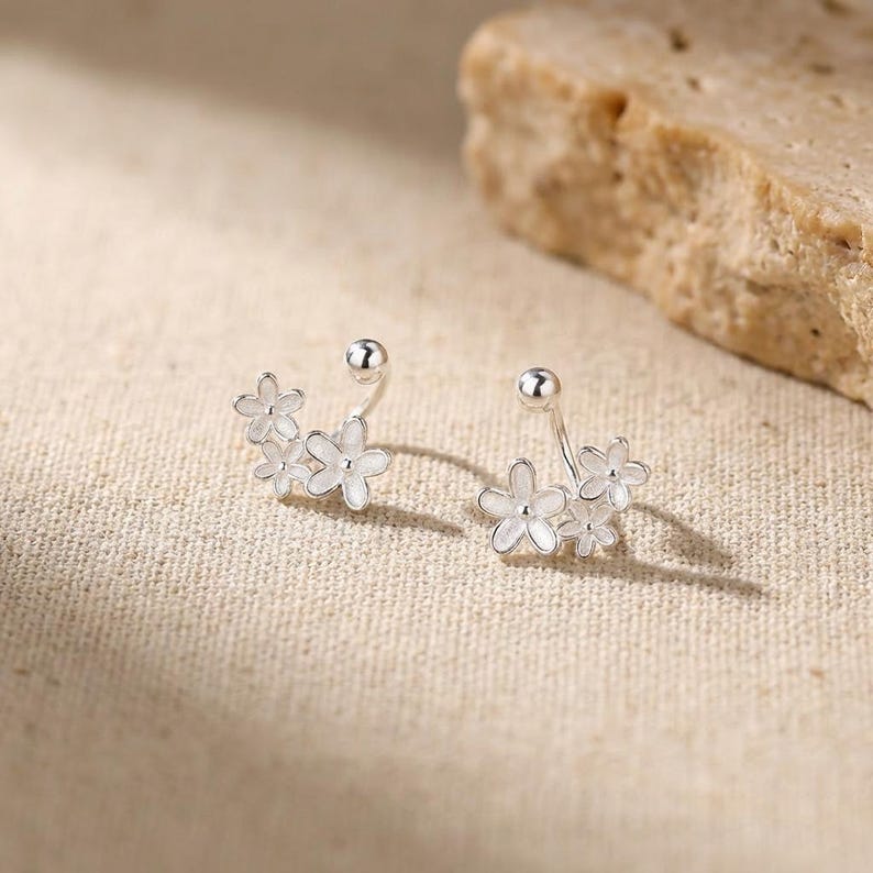 Op de afbeelding: Een paar zilverkleurige bloemvormige oorbellen. Elke oorbel heeft een cluster van kleine, delicate bloemen met witte bloemblaadjes. De oorbellen hebben een post-back ontwerp voor doorboorde oren. De oorbellen worden gepresenteerd op een neutrale ondergrond.