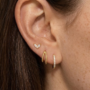 May include: Close-up of an ear adorned with three gold earrings. The top earring is a small, gold stud with a marquise-shaped, clear stone. Below it are two gold hoop earrings, one twisted and the other encrusted with small, clear stones.