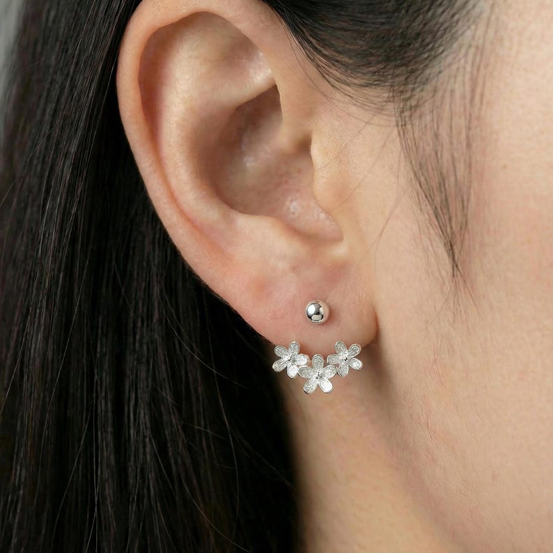 Op de afbeelding: Zilveren bloemen oorbellen met een knop en een cluster van drie bloemen. De oorbellen worden op het oor gedragen, met de knop in de bovenste oorlel en het bloemendesign eronder. Een delicaat en vrouwelijk accessoire.