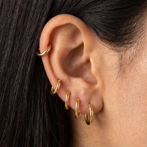 May include: A close-up of an ear adorned with multiple gold hoop earrings of varying sizes. The earrings are a bright, shiny gold color and are arranged in a stylish, layered design. The ear is set against a backdrop of dark hair.