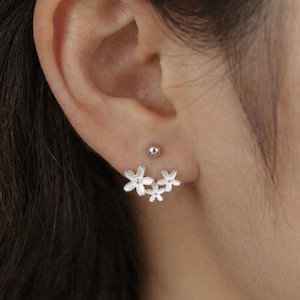 Op de afbeelding: Zilverkleurige bloemen oorbellen. De oorbellen hebben een cluster van drie kleine bloemen, met een kleine zilveren stud erboven. De oorbellen worden op een oor gedragen.