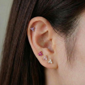 May include: Close-up of an ear with multiple piercings. The piercings feature small, gold-coloured stud earrings. Each earring has a cluster of gemstones in shades of purple, pink, and clear. The ear is set against a blurred background.