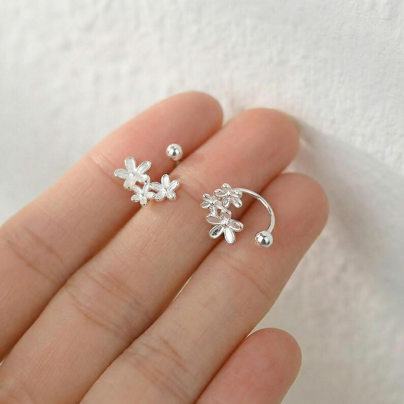 Op de afbeelding: Zilverkleurige bloemen oorbellen. E&eacute;n oorbel heeft een cluster van drie kleine bloemen, de andere een gebogen ontwerp met een bloemcluster en een kleine bal. Er is ook een kleine oorbel aanwezig.