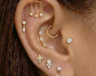 Op de afbeelding: Een oor versierd met meerdere goudkleurige oorbellen. De oorbellen omvatten een bungelende staaf met drie bedels, een ring, studs en diverse oorhangers met heldere edelstenen. De sieraden zijn op het oor gerangschikt en tonen verschillende stijlen.