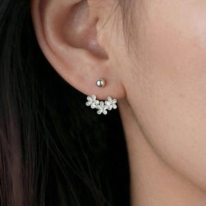 Op de afbeelding: Een paar zilverkleurige oorbellen. E&eacute;n is een kleine, ronde knop. De andere heeft een cluster van vijf kleine, bloemvormige bedels met heldere, sprankelende accenten. De oorbellen worden op een oor gedragen.