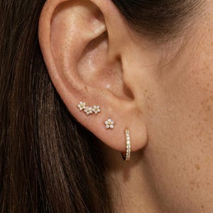 May include: Close-up of an ear with several gold earrings. The earrings include a floral cluster, a single flower stud, and a small hoop. The earrings are decorated with tiny, sparkling stones.