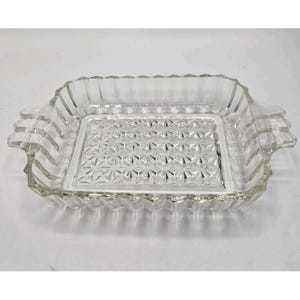 May include: Clear glass rectangular serving dish with a textured geometric pattern on the base. The dish has a scalloped edge and handles on each side. The glass is transparent, allowing light to pass through.