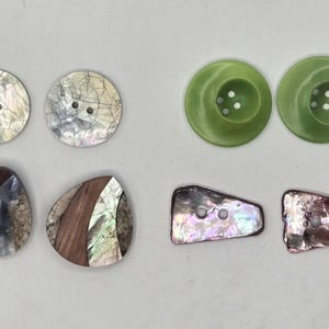 May include: A collection of decorative buttons in various shapes and materials. Includes two round, iridescent shell buttons, two green, circular buttons, and two irregularly shaped buttons with wood and shell accents.