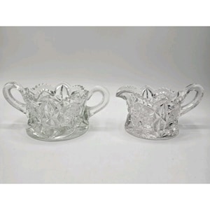 May include: A clear glass sugar bowl and creamer set. The sugar bowl has two handles and a scalloped rim. The creamer has a spout and a handle. Both pieces have a starburst pattern.