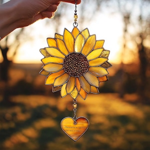 Puede incluir: Atrapasoles de girasol de vidrieras con pétalos amarillos y dorados y un centro marrón. Un dije en forma de corazón con texto cuelga debajo. El atrapasoles está suspendido por una cadena.