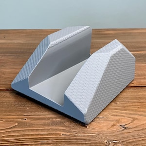 May include: A gray, geometric phone stand with a textured surface. The stand has a U-shaped design with angled sides and a smooth interior. The stand is made of a light gray material and sits on a wooden surface.
