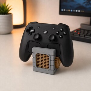 May include: A black wireless game controller resting on a gray, block-shaped stand. The stand features a tan, grid-patterned center panel and a visible tan gear on the side. The controller has a central Steam logo, two joysticks, and standard buttons.