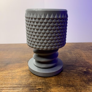 May include: A gray, 3D-printed decorative cup with a textured, diamond-patterned exterior. The cup sits atop a tiered base, creating a unique, modern design. The cup is approximately 4 inches tall.