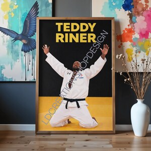 Peut inclure: Impression encadrée de Teddy Riner en judogi blanc et ceinture noire, à genoux, les bras levés. Le texte "TEDDY RINER" en lettres dorées se détache sur fond noir, avec une bande jaune en bas.