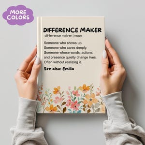 Puede incluir: Un cuaderno con las palabras "DIFFERENCE MAKER" y su definición impresas en la portada. La portada también presenta un diseño floral con flores naranjas, rosas y amarillas. Las palabras "MORE COLORS" están en una burbuja morada.