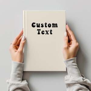 May include: A cream-colored notebook with the words "Custom Text" in bold, black letters. The notebook is held by two hands with manicured nails, against a light gray background. The notebook appears to be blank inside.