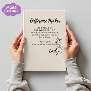 Puede incluir: Un diario de color crema con las palabras "Difference Maker" y una cita inspiradora impresa en negro. El diario está sostenido por dos manos. El texto incluye el nombre "Emily" y una ilustración floral.