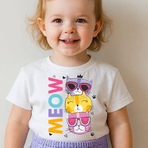 May include: White t-shirt with a colourful design of three cartoon cats wearing sunglasses, stacked on top of each other. The word "MEOW" is printed vertically. Small paw prints and hearts are scattered around.