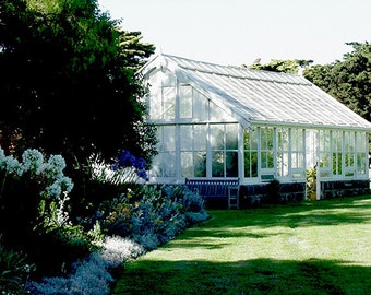 May include: A white greenhouse with a glass roof and windows. The greenhouse is surrounded by a lush green lawn and a garden with white and blue flowers.