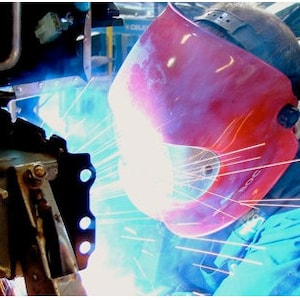 Könnte beinhalten: Ein Schweißer mit einem roten Schweißhelm mit klarem Visier und einem blauen Overall schweißt Metall. Funken fliegen aus dem Schweißbrenner.