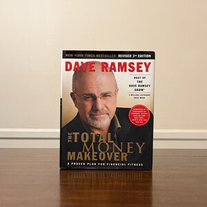 Op de afbeelding: De omslag van "The Total Money Makeover" van Dave Ramsey. Het boek is een New York Times-bestseller, herziene 3e editie. De omslag toont een foto van Dave Ramsey en de titel in grote, vette letters. Het boek ligt op een houten oppervlak.