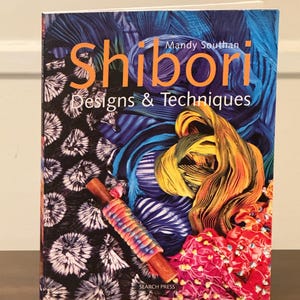 May include: A book titled "Shibori Designs & Techniques" by Mandy Southan. The cover features colorful textiles in shades of blue, yellow, and red, along with a wooden tool. The book is published by Search Press.