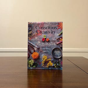 Może przedstawiać: Książka zatytułowana "Conscious Creativity" z podtytułem "The Workbook: experiment, explore, create". Okładka przedstawia kolaż materiałów artystycznych, w tym przędzy, kredek i kwiatów. Książka leży na ciemnobrązowym drewnianym stole.