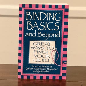 Puede incluir: Un libro titulado "BINDING BASICS and Beyond" con una cubierta azul marino y un borde a rayas rosa y azul. El libro presenta el texto "GREAT WAYS TO FINISH YOUR QUILT" y es de los editores de Quilter's Newsletter Magazine y Quiltmaker.