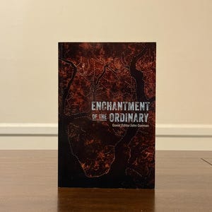 May include: A hardcover book titled "Enchantment of the Ordinary" with a dark red and brown cover design. The cover features a textured pattern and the text is in white. The book is standing upright on a wooden surface.