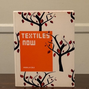 Op de afbeelding: Een boek getiteld "TEXTILES NOW" door Drusilla Cole. De omslag heeft een crèmekleurige achtergrond met een patroon van zwarte boom silhouetten met rode bladeren. De titel staat in witte blokletters op een oranje rechthoek.