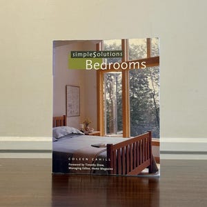 May include: A softcover book titled simpleSolutions Bedrooms by Coleen Cahill with a foreword by Timothy Drew. The cover features a photograph of a bedroom with a wooden bed frame, a grey bedspread, and large windows looking out onto trees.