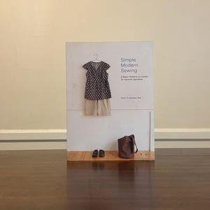 May include: A softcover book titled Simple Modern Sewing: 8 Basic Patterns to Create 25 Favorite Garments by Shufu To Seikatsu Sha. The cover features a dark patterned top on a hanger, beige shorts, a brown bag, and dark shoes on a floor.
