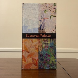Puede incluir: Un libro de tapa dura titulado "Seasonal Palette" con una cubierta colorida. La cubierta está dividida en secciones con patrones florales y geométricos, y una ilustración de garza. El libro está de pie sobre una superficie de madera.