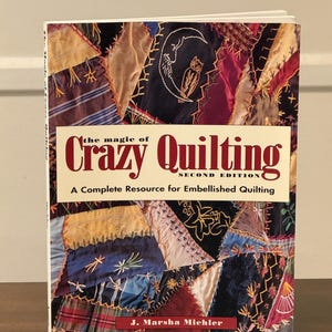 Peut inclure: Un livre intitulé "the magic of Crazy Quilting, Second Edition" avec un motif de patchwork sur la couverture. La couverture présente le texte "A Complete Resource for Embellished Quilting" et le nom de l'auteur, J. Marsha Michler.