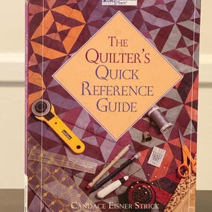 Puede incluir: Un libro titulado "The Quilter's Quick Reference Guide" de Candace Eisner Strick. La portada presenta herramientas de acolchado como un cortador rotatorio, tijeras y agujas. El fondo tiene un patrón de edredón geométrico en tonos morado, naranja y marrón.