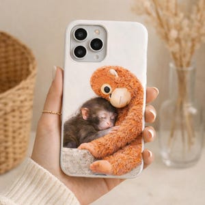 May include: White phone case featuring a heartwarming illustration of a sleeping baby monkey being embraced by a plush orange monkey. The case has a glossy finish and a camera cutout.