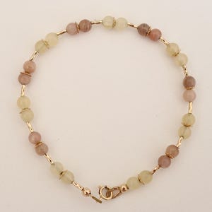 May include: A beaded bracelet featuring alternating light green and pinkish-brown gemstone beads. The beads are strung on gold-colored metal bars and connected with a gold-colored clasp. The bracelet is designed for a delicate and feminine look.