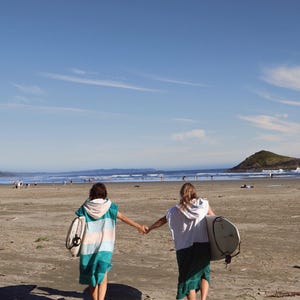 Puede incluir: Dos personas caminando por una playa arenosa hacia el océano, tomadas de la mano. Una lleva una toalla con capucha a rayas turquesa y blanca, la otra una parte superior con capucha blanca y una falda verde. Ambos llevan tablas de surf.