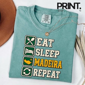 Puede incluir: Una camiseta de manga corta color verde espuma de mar con un gráfico de cuatro cuadrados apilados. Los cuadrados contienen iconos de comer, dormir, un mapa de Madeira y un símbolo de reciclaje, junto al texto EAT, SLEEP, MADEIRA, REPEAT.