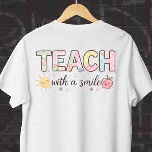 Pode incluir: Uma t-shirt branca com a palavra TEACH em letras de bloco com padrões coloridos em tons pastel. Abaixo do texto, a frase with a smile está escrita em letra cursiva preta, acompanhada por um pequeno sol amarelo e uma maçã rosa com rostos.