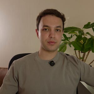Könnte beinhalten: Eine Person trägt ein graues Rundhals-Sweatshirt mit einem kleinen schwarzen quadratischen Mikrofon am Kragen. Der Hintergrund umfasst eine Pflanze mit grünen Blättern und eine neutralfarbene Wand.