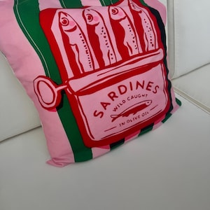 Puede incluir: Cojín decorativo rosa con un diseño de lata de sardinas. El cojín presenta un fondo a rayas rosas y verdes con una ilustración roja de una lata de sardinas. La lata está etiquetada como "Sardines Wild Caught in Olive Oil."
