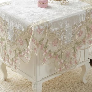 Puede incluir: Una mesita de noche blanca con un mantel bordado decorativo con motivos florales rosas y verdes. Una pequeña caja de joyas rosa y una figura de carroza dorada están encima. La mesa está sobre una alfombra color crema.